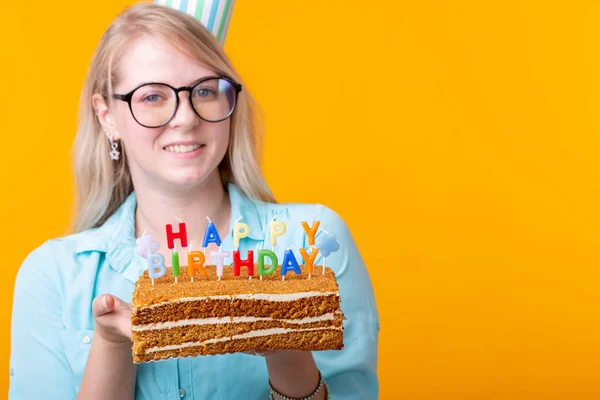 キャップと自家製ケーキを手に持つポジティブな面白い若い女性は、コピースペースと黄色の背景にポーズ。記念日と誕生日のコンセプト. — ストック写真