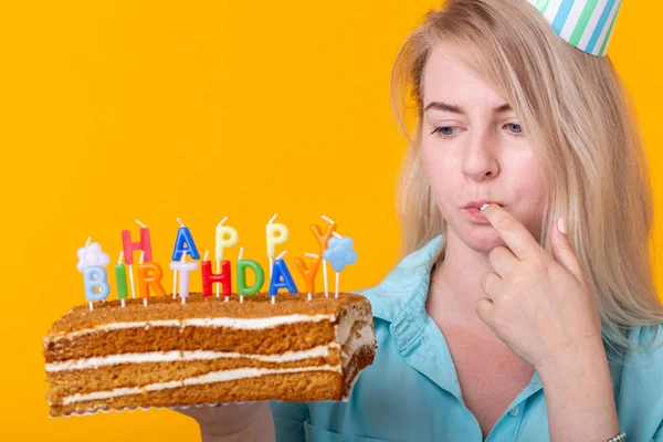 Közeli fel őrült vidám fiatal nő a papír gratuláló kalap gazdaság sütemények boldog születésnapot állva egy sárga háttér. Jubileumi gratuláció koncepció. — Stock Fotó