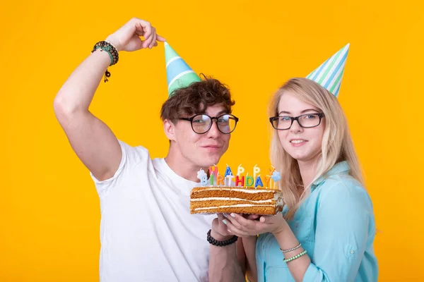 Jovem alegre casal charmoso cara e menina bonito em chapéus de papel fazer cara tola e segurar em suas mãos um bolo com a inscrição aniversário de pé sobre um fundo amarelo. Conceito saudações e — Fotografia de Stock