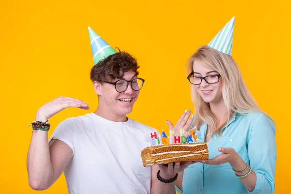 Young cheerful students charming girl and nice guy in greeting paper caps holding a cake. Concept of congratulations on the birthday and anniversary