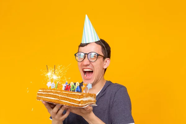 Divertente ragazzo positivo in occhiali tiene tra le mani una torta fatta in casa con l'iscrizione buon compleanno in posa su uno sfondo giallo. Concetto di vacanze e anniversari . — Foto Stock
