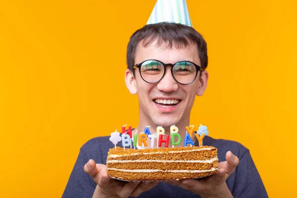 Смешной позитивный парень в очках держит в руках домашний торт с надписью "С днем рождения, позируя на жёлтом фоне". Концепция праздников и юбилеев . — стоковое фото