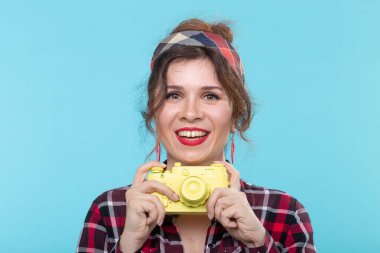 Mavi bir arka plan karşı sarı bir vintage kamera ile fotoğraf çekmek ekose gömlek giyen olumlu bir güzel genç kadının Closeup portre. Konsept hobiler ve fotoğraflar