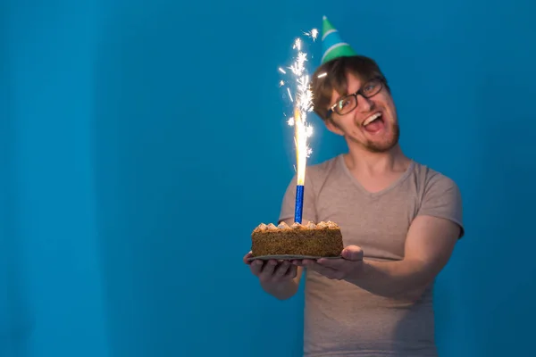 Crazy cheerful guy student in glasses greeting with a paper hat holding a cake with burning fireworks in his hands. Holiday birthday concept — Stock Photo, Image