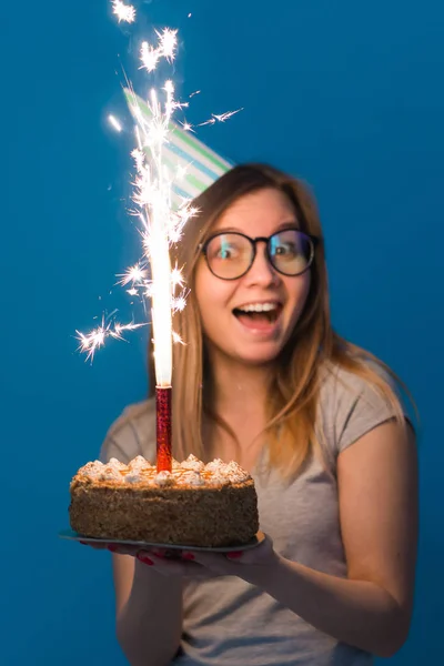 Jovem estudante menina borrada alegre em óculos segurando um bolo de parabéns com uma vela em pé sobre um fundo azul. Conceito de aniversário . — Fotografia de Stock