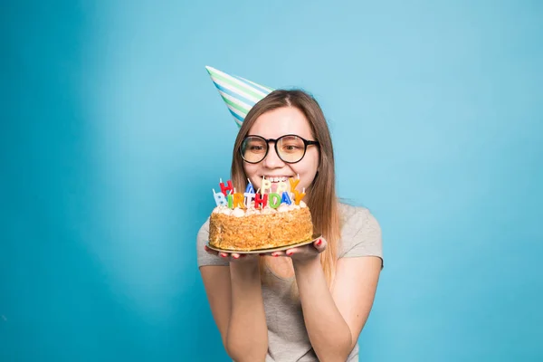 Очаровательная юная сумасшедшая студентка в бумажной шляпе с праздничным тортом в руках, стоящая на синем фоне с копировальным местом. Рекламное пространство . — стоковое фото