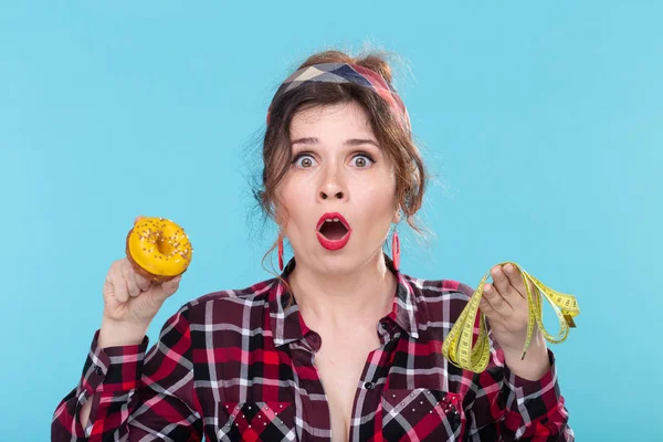 Close-up shocked surprised young beautiful woman looking at measuring tape and holding a green donut in hands posing on a blue background. Concept of diet and rejection of harmful high-calorie foods.