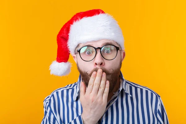 Verraste jongeman, de Kerstman, sluit zijn mond terwijl hij naar de camera kijkt op een gele achtergrond. Het concept van felicitaties voor het nieuwe jaar en Kerstmis. Advertentieruimte. — Stockfoto