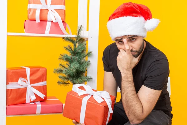 Joven hombre guapo en un sombrero de Santa Claus está sosteniendo regalos de felicitación al lado de un árbol de Navidad. El concepto de las fiestas navideñas y un nuevo año . — Foto de Stock