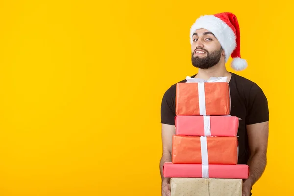 Ontevreden jonge man met een baard in een kerstman hoed bevat vijf geschenkdozen poseren op een gele achtergrond met copyspace. Concept van cadeaus en groeten voor Kerstmis en Nieuwjaar. — Stockfoto