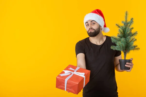 Przystojny młody człowiek trzyma czerwony prezent pudełko i Choinka w rękach stwarzających w nowym roku Cap na żółtym tle z copyspace. Wesołych Świąt Bożego Narodzenia i szczęśliwego nowego roku koncepcja pozdrowienia. — Zdjęcie stockowe