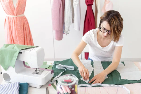 La joven diseñadora hace marcas para un nuevo producto de costura sentado en la mesa junto a la máquina de coser. Concepto de negocio creativo y diseño . — Foto de Stock