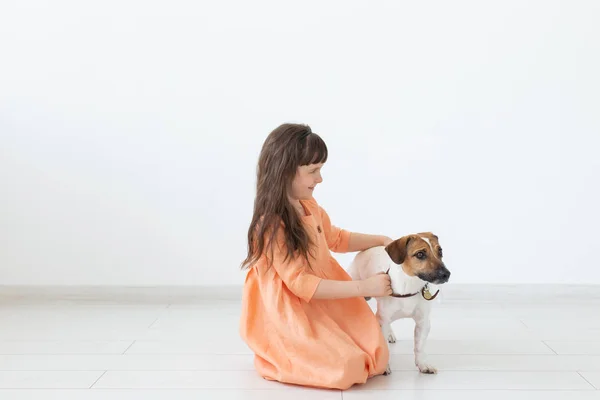 어두운 머리를 가진 어린 소녀는 흰색 벽의 배경에 복숭아 드레스바닥에 앉아있는 동안 그녀의 사랑하는 개와 함께 재생됩니다. 어린이와 동물을 돌보는 개념. 광고 공간. — 스톡 사진