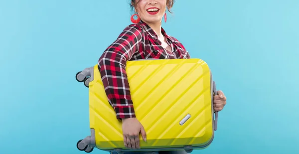 Θετική νεαρή χαμογελαστή γυναίκα με καρό πουκάμισο κρατώντας μια κίτρινη βαλίτσα που ποζάρει σε μπλε φόντο. Έννοια του τουρισμού και των ταξιδιών — Φωτογραφία Αρχείου