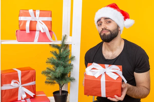 Jovem bonitão de chapéu de Papai Noel está segurando presentes de parabéns ao lado de uma árvore de Natal. Conceito de férias de Natal e Ano Novo . — Fotografia de Stock