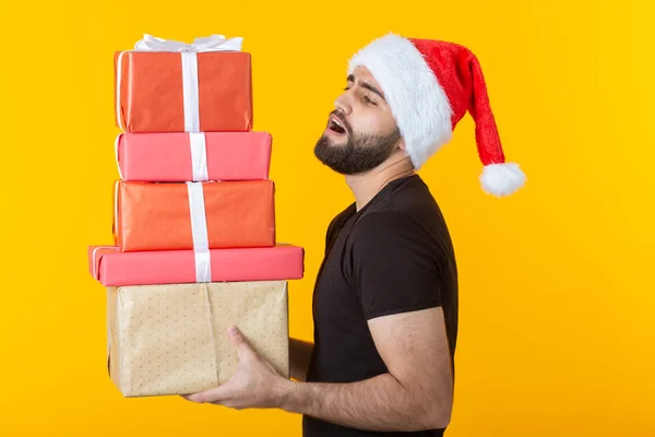 Ontevreden jonge man met een baard in een kerstman hoed bevat vijf geschenkdozen poseren op een gele achtergrond. Concept van cadeaus en groeten voor Kerstmis en Nieuwjaar. — Stockfoto