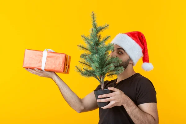 Przystojny młody człowiek trzyma czerwony prezent pudełko i Choinka w rękach stwarzających w nowym roku Cap na żółtym tle. Wesołych Świąt Bożego Narodzenia i szczęśliwego nowego roku koncepcja pozdrowienia. — Zdjęcie stockowe