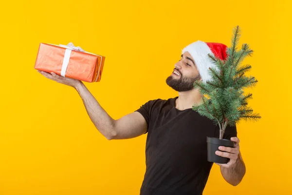 Joven guapo sosteniendo una caja de regalo roja y un árbol de Navidad en sus manos posando en una gorra de Año Nuevo sobre un fondo amarillo. Feliz Navidad y Feliz Año Nuevo concepto de felicitaciones . —  Fotos de Stock