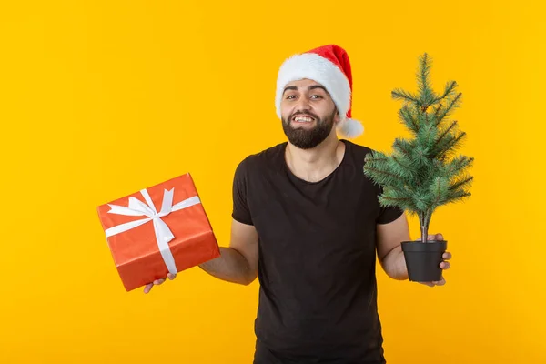 Knappe jonge man met een rode geschenk doos en kerstboom in zijn handen poseren in een nieuwe jaar GLB op een gele achtergrond. Merry Christmas en Happy New Year groeten concept. — Stockfoto