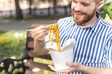 Güneşli bir yaz gününde bir park bankında otururken bir öğle yemeği kutusundan Çin erişte yiyen genç yakışıklı hipster adam Yan görünümü. Sağlıklı ve besleyici bir atıştırmalık kavramı.