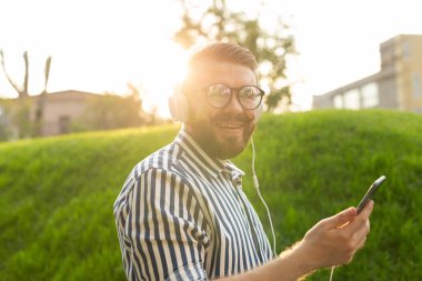 Pozitif genç erkek hipster güneşli sıcak bir yaz gününde parkta akıllı telefon kulaklık ve yüksek hızlı kablosuz internet kullanarak favori online radyo dinler.
