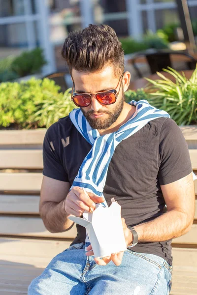 Joven hipster masculino con bigote y barba está comiendo fideos chinos con palillos de madera de una lonchera blanca. concepto de cocina asiática . — Foto de Stock