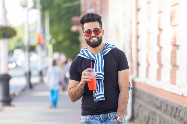 Retrato de un joven árabe alegre positivo con un vaso de jugo con una pajita mientras camina por la ciudad en un cálido día soleado de verano. El concepto de descanso después del estudio y el trabajo los fines de semana . — Foto de Stock