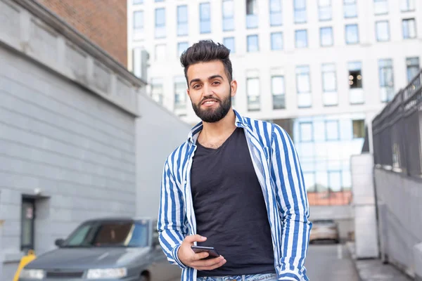 Jeune homme hipster arabe élégant avec barbe bavarder dans les réseaux sociaux en utilisant un smartphone et Internet sans fil par une chaude journée d'été . — Photo