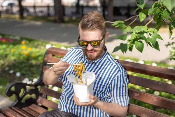 Pohled na mladého hezkého hipíky, který Konzumujte čínské nudle z krabice na oběd, když sedí na lavičce v parku za slunného letního dne. Koncepce zdravého a výživných svačinka. — Stock fotografie