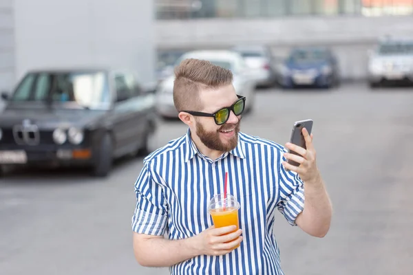 Vista lateral de un joven y elegante hombre hipster con gafas y un bigote y jugo en sus manos charlando en las redes sociales usando un teléfono inteligente e internet inalámbrico en un cálido día de verano . — Foto de Stock