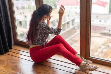 Hayal kurma, düşüncelilik ve yalnızlık konsepti - Genç beyaz kadın oturma odasında oturuyor ve büyük pencereden dışarı bakıyor