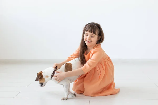 Pequeña chica encantadora con el pelo oscuro en un vestido largo está sentado al lado del perro Jack Russell Terrier en el suelo de su habitación. El concepto de inocencia infantil y cuidado infantil. Espacio de copia . — Foto de Stock