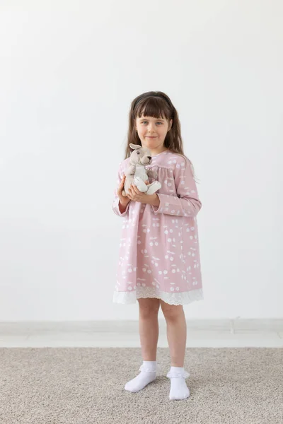 Beautiful little girl holding a teddy rabbit in a pink dress next to a white wall. The concept of cute children and childrens toys and clothing. — Stock Photo, Image