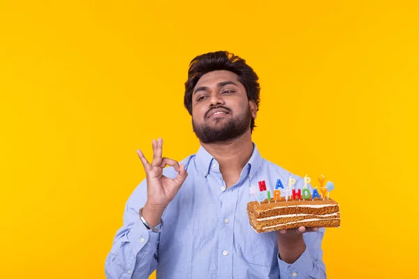 Positivo engraçado jovem indiano com um boné e um bolo caseiro em suas mãos posando em um fundo amarelo. Aniversário e conceito de aniversário . — Fotografia de Stock