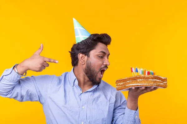 Pazzo allegro giovane indiano in carta cappello di congratulazione che tiene torte buon compleanno in piedi su uno sfondo giallo. Concetto di congratulazioni giubilari . — Foto Stock