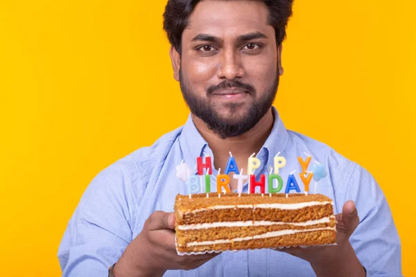 Close-up of crazy cheerful young indian man in paper congratulatory hat holding cakes happy birthday standing on a yellow background. Jubilee congratulations concept.