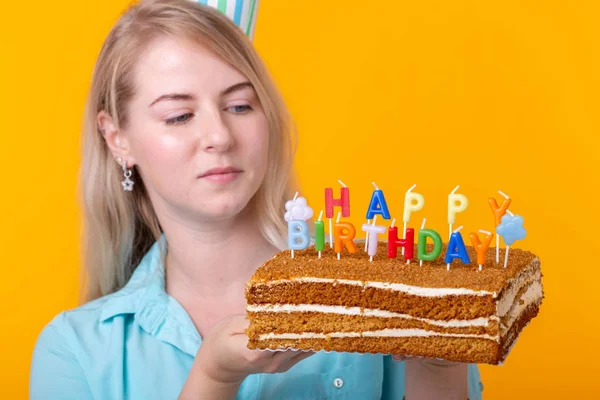 Close-up de mulher jovem alegre em papel chapéu congratulatório que mantém bolos aniversário feliz em pé em um fundo amarelo. Conceito de parabéns Jubileu . — Fotografia de Stock