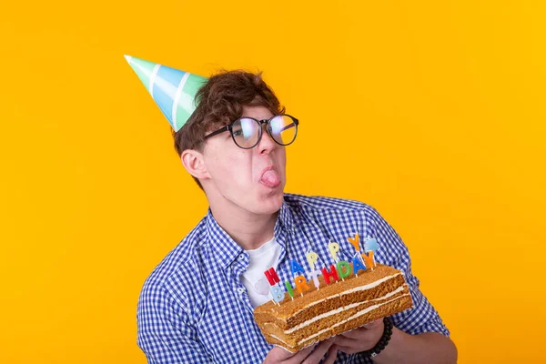 Divertente ragazzo positivo tiene in mano una torta fatta in casa con l'iscrizione buon compleanno in posa su uno sfondo giallo. Concetto di vacanze e anniversari . — Foto Stock
