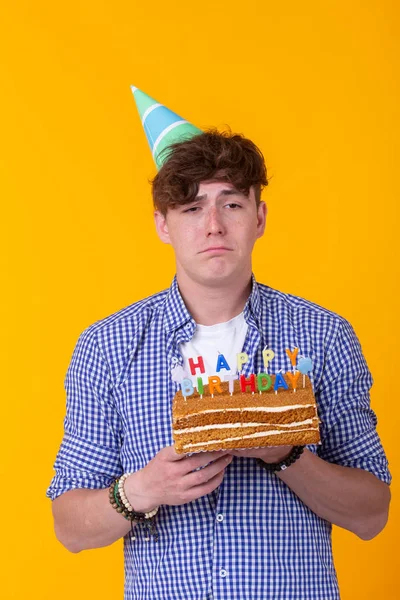 Crazy vidám fiatalember papíron gratuláló kalap gazdaság sütemények boldog születésnapot állva egy sárga háttér. Jubileumi gratuláció koncepció. — Stock Fotó