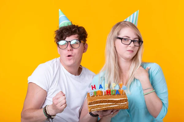 Engraçado nerd homem e mulher estão usando feriados bonés e óculos segurando bolo de aniversário com velas sobre fundo amarelo — Fotografia de Stock