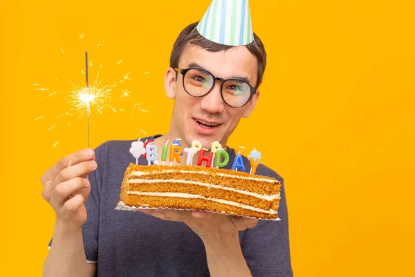 Позитивний смішний молодий азіатський хлопець з шапочкою і палаючою свічкою і домашній торт в руках позує на жовтому тлі. Концепція ювілею та дня народження . — стокове фото