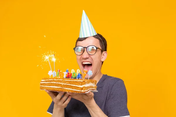 Loco alegre chico asiático joven con gafas sosteniendo una vela ardiente en sus manos y un pastel casero de felicitación sobre un fondo amarillo. Concepto de celebración de cumpleaños y aniversario . — Foto de Stock