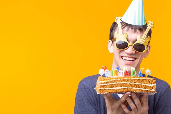 Jovem alegre louco em óculos e chapéus de papel parabéns segurando bolos feliz aniversário de pé em um fundo amarelo com espaço de cópia. Conceito de parabéns Jubileu . — Fotografia de Stock