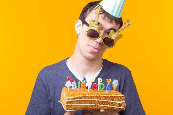 Retrato de um cara positivo engraçado com um boné de papel e óculos segurando um bolo caseiro congratulatório em suas mãos em um fundo amarelo. Conceito e diversão e celebração . — Fotografia de Stock