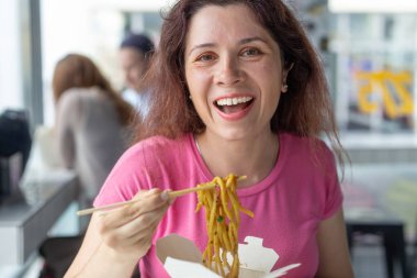 Bir kafede Çin eriştesi yiyen ve pencereden dışarı bakan genç ve neşeli bir kızın portresi. Sağlıklı Asya mutfağı kavramı.