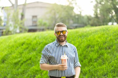 Sakallı, süt sallama lı ve sıcak bir yaz gününde parkta yürüyen şehrin manzarasına hayran olan şık bir genç adamın yan görünümü. Dinlenme ve rahatlama kavramı.