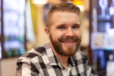 Bir kafede poz veren bıyıklı ve sakallı sevimli pozitif genç hipster adam. Komik neşeli erkekler kavramı.