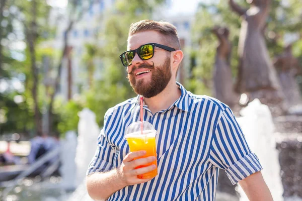 Lindo hipster joven y elegante con bigote y barba y gafas bebiendo jugo con una pajita mientras está sentado en un parque de la ciudad sobre el fondo de una fuente. Concepto vacaciones de verano . — Foto de Stock