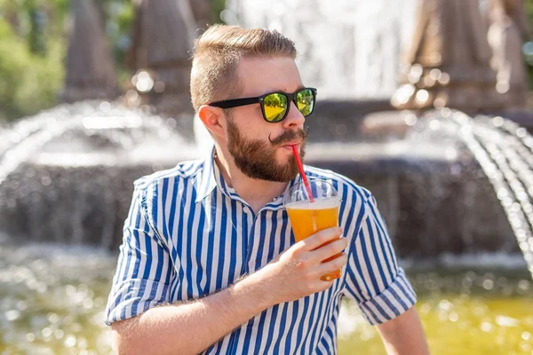 Lindo hipster joven y elegante con bigote y barba y gafas bebiendo jugo con una pajita mientras está sentado en un parque de la ciudad sobre el fondo de una fuente. Concepto vacaciones de verano . — Foto de Stock