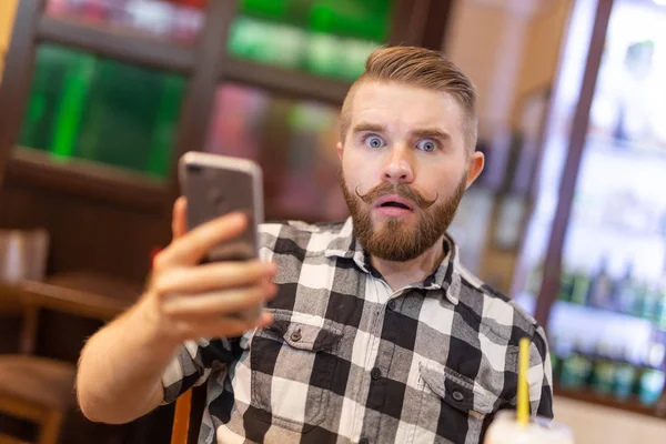 Потрясенный молодой хипстер пьет какао из соломы и смотрит на сообщения в социальных сетях, сидя за столом в кафе. Концепция чата и социальных сетей . — стоковое фото
